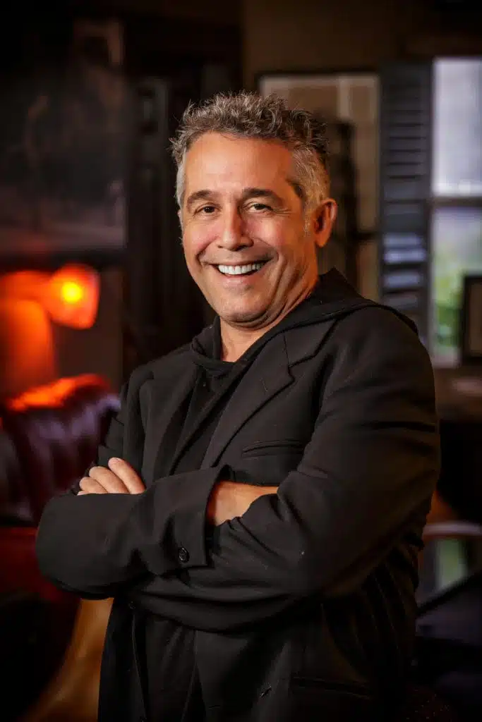 A man with short, wavy gray hair smiles with arms crossed, wearing a black jacket and shirt. He stands indoors in a warmly lit room with dark furniture and a glowing lamp in the background, presenting a professional business headshot atmosphere.