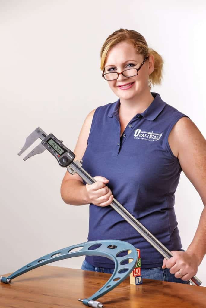 A woman with glasses holds a large caliper and smiles at the camera in this creative business headshot. She wears a navy sleeveless shirt and stands behind a table with a blue measuring tool and colorful wooden blocks.