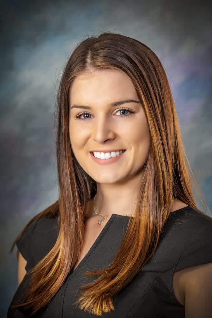 Smiling woman with straight brown hair, wearing a dark short-sleeve top and a necklace, posed in front of a blurred, multicolored studio background—an ideal business headshot capturing both professionalism and warmth.