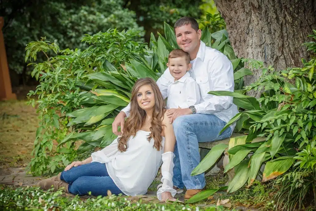 A family of three, all in white tops and jeans, poses happily outdoors by a large tree and green plants. The mother is sitting on the ground, while the father and young boy are seated on a bench. Captured by Kirk Voclain Photography, this moment perfectly reflects the warm style of a Houma photographer.