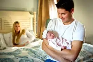 A man in a white t-shirt cradles a newborn baby wrapped in a blanket, while a woman sits on a bed in the background, smiling and watching them. The room is softly lit and cozy, perfectly capturing the warmth of family moments often seen in Kirk Voclain Photography, your trusted Houma photographer.