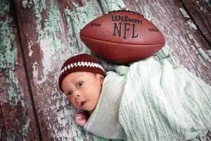A baby wrapped in a green blanket lies on a wooden floor next to an NFL football. The baby wears a knitted brown and white hat that resembles a football. Captured by Houma photographer Kirk Voclain Photography, this charming scene beautifully blends warmth and sporty spirit.