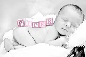 A newborn baby sleeps on a soft blanket with pink wooden blocks spelling PIPER resting on their side. Captured by Kirk Voclain Photography, this black and white photo highlights the gentle moment, with the pink blocks providing a charming pop of color. Perfect for anyone seeking a Houma photographer who creates timeless memories.