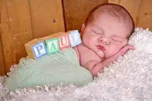 A sleeping newborn wrapped in a light green blanket, lying on a soft white surface, with wooden blocks spelling PAUL resting on their side—captured with the artistry of Kirk Voclain Photography, a renowned Houma photographer.