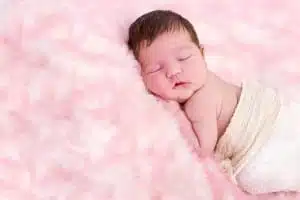 A newborn baby wrapped in a soft white blanket sleeps peacefully on a fluffy, pale pink surface, beautifully captured by Kirk Voclain Photography, a renowned Houma photographer.