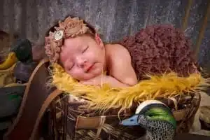 A sleeping baby dressed in a brown ruffled outfit and headband lies on a yellow fuzzy blanket, surrounded by duck decoys—a heartwarming moment beautifully captured by Houma photographer Kirk Voclain Photography.