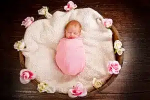 A newborn baby wrapped in a pink blanket sleeps on a soft white blanket in a round wooden basket, surrounded by pink and white flowers on a dark wooden floor. The peaceful newborn scene is the epitome of innocence and serenity.