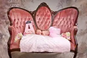A newborn baby in a pink tutu sleeps on a vintage pink sofa, surrounded by flowers and a pink decorative letter A on a soft, embroidered blanket, beautifully captured by Kirk Voclain Photography, your trusted Houma photographer.