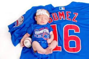 A baby wearing a Chicago Cubs World Series Champions onesie and white headband sleeps on a large Cubs jersey, next to a baseball, in this adorable moment captured by Kirk Voclain Photography, your trusted Houma photographer.