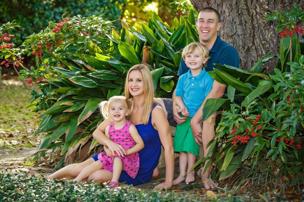 A smiling family of four poses outdoors by a large tree, surrounded by green plants and red berries—capturing the warmth and joy often seen in family portraits. The mother and daughter sit in front, while the father and son kneel behind them, all wearing colorful, casual clothing.