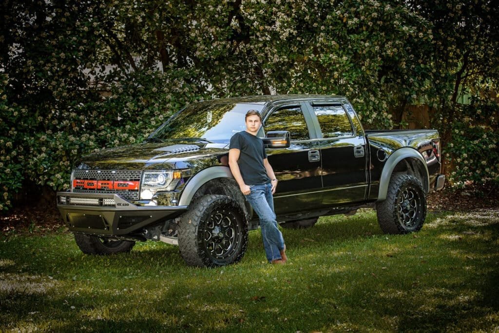 A person in a black t-shirt and jeans leans against a large black Ford pickup truck parked on grass, with trees and greenery in the background—an ideal setting for senior portraits Houma LA that capture both personality and style.