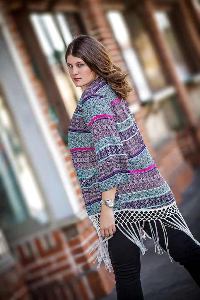 A woman with wavy brown hair wears a patterned shawl with fringe, black pants, and a watch, standing outdoors near a brick building—one of the best senior picture locations in Thibodaux, LA—while looking over her shoulder.