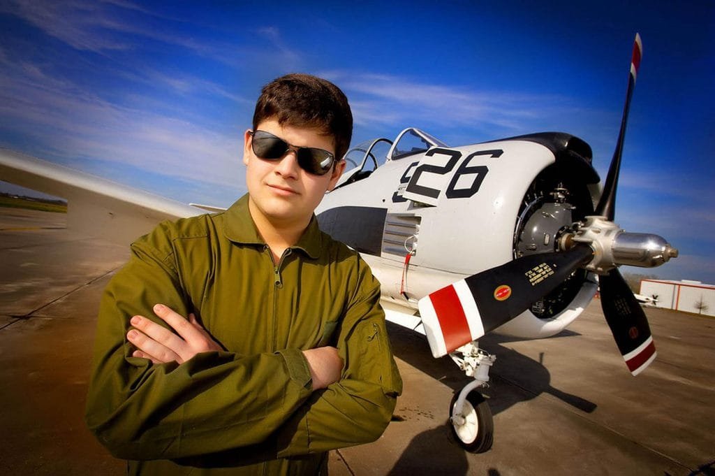 A young man in sunglasses and a green jacket stands confidently with arms crossed in front of a vintage propeller airplane marked with the number 26 on a sunny day—one of the best senior picture locations in Thibodaux, LA for capturing unique and memorable portraits.
