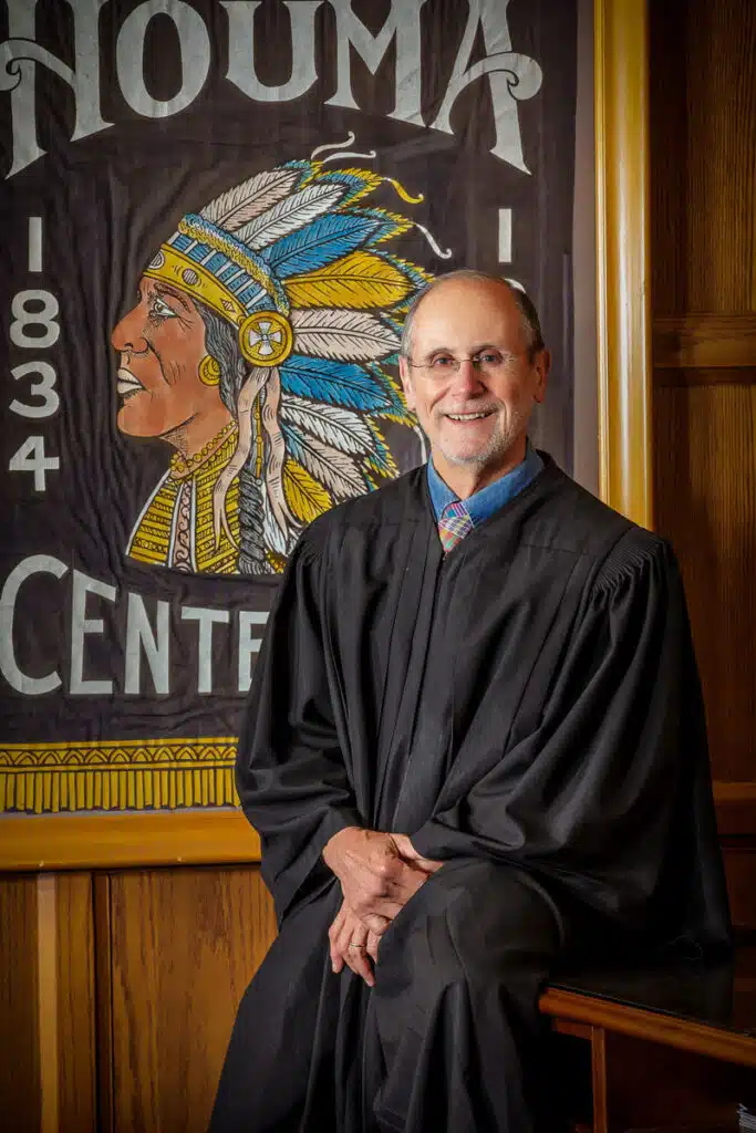A judge in a black robe smiles while seated in front of a banner featuring a Native American chief in a feathered headdress and the text HOUMA and 1834 CENTER, captured with the skillful touch of a headshot photographer Houma.