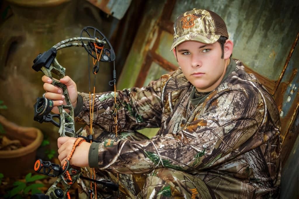 03-11 A young person in camouflage gear and cap sits outdoors, holding a compound bow, with greenery and rustic metal structures in the background—a perfect setting for senior pictures with cars, trucks, hunting and fishing themes.