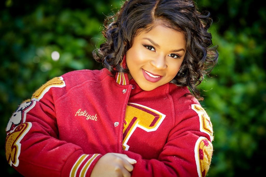 A smiling young woman with styled dark hair wears a red varsity jacket with the name Aalyah embroidered on it, standing outdoors in front of green foliage—one of the best senior picture locations in Thibodaux, LA.