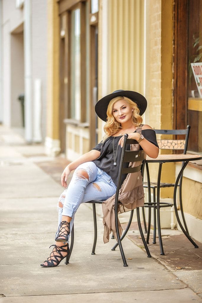 A woman with blonde hair in a black hat, black top, ripped jeans, and strappy heels sits confidently at an outdoor café table on a sidewalk—one of the best senior picture locations in Thibodaux, LA—looking at the camera and smiling slightly.