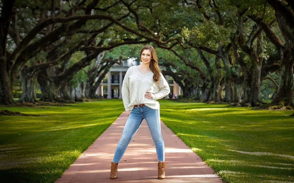 A woman in jeans and a sweater stands smiling on a red brick walkway, lined with large oak trees, leading to a white house in the background on a bright day—one of the best senior picture locations in Thibodaux, LA.