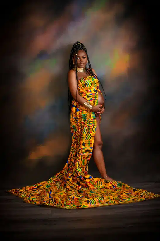Pregnant woman in a vibrant, patterned dress stands barefoot on a wooden floor, holding her belly, with a flowing train and a colorful, blurred backdrop behind her—an elegant moment beautifully captured by a skilled maternity photographer Houma. Kirk Voclain Photography