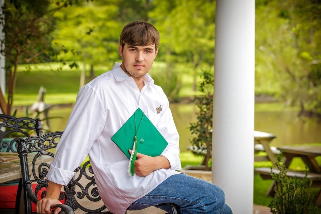 A young man in a white shirt and jeans sits outdoors at one of the best senior picture locations in Thibodaux, LA, holding a green graduation cap and gown. A pond, trees, and picnic tables are in the background.