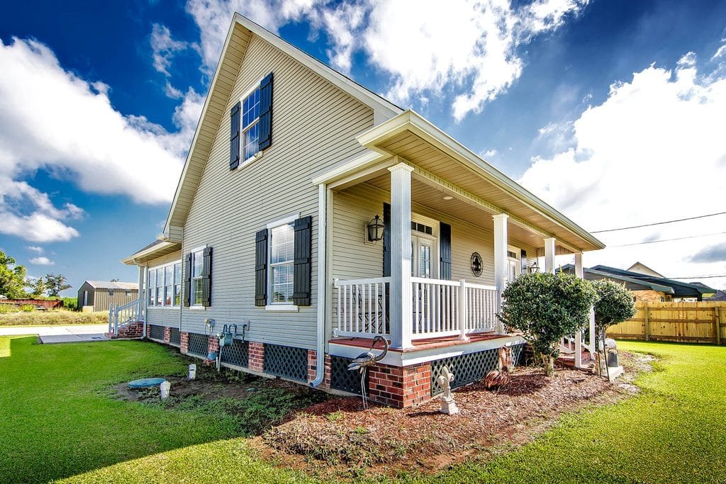 A beige house with black shutters, a covered front porch, brick foundation, and white railings sits on a green lawn under a blue sky with clouds, providing an excellent example of realtor photography.