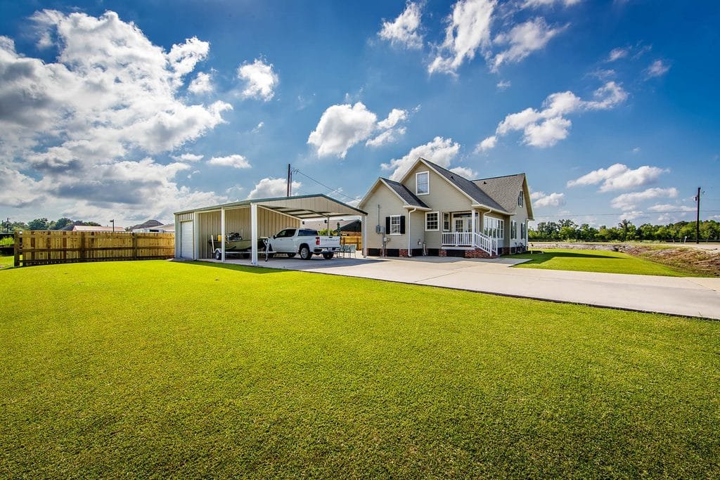 A beige house with a covered carport, two vehicles parked, and a large well-kept lawn under a partly cloudy blue sky—this image serves as one of the standout realtor photography examples. A wooden fence and trees are visible in the background.