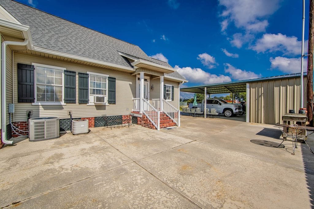 A beige house with white trim, black shutters, and small front steps overlooks a large concrete driveway—an ideal setting highlighted in many realtor photography examples. A metal carport with vehicles is visible in the background, under a partly cloudy sky.
