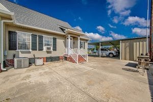 A beige house with white trim, black shutters, and small front steps overlooks a large concrete driveway—an ideal setting highlighted in many realtor photography examples. A metal carport with vehicles is visible in the background, under a partly cloudy sky.