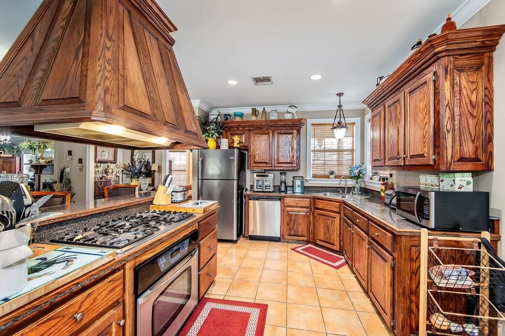 A spacious kitchen with wooden cabinets, stainless steel appliances, a gas stove, tiled floor, and red rugs. Countertops hold small appliances, plants, and decor. Windows let in natural light—an inviting space showcased in many realtor photography examples.