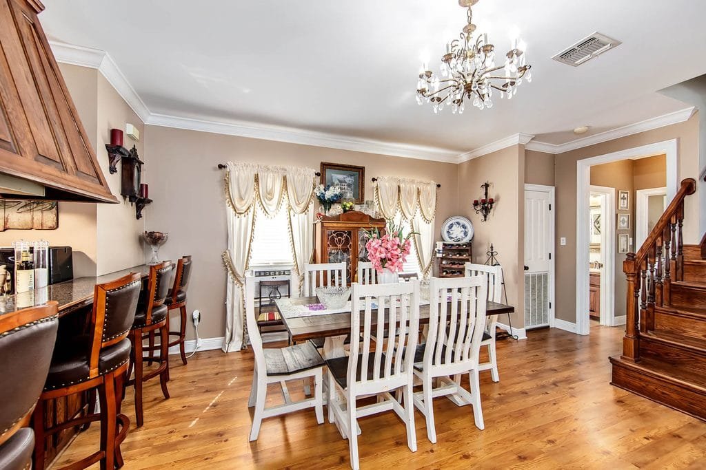 Bright dining room with a wooden table, white chairs, flowers centerpiece, and barstools at a kitchen counter. Elegant curtains, a chandelier, and wooden floors add warmth. A staircase is visible to the right—an inviting space that showcases realtor photography examples at their finest.