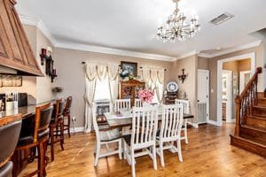 Bright dining room with a wooden table, white chairs, flowers centerpiece, and barstools at a kitchen counter. Elegant curtains, a chandelier, and wooden floors add warmth. A staircase is visible to the right—an inviting space that showcases realtor photography examples at their finest.