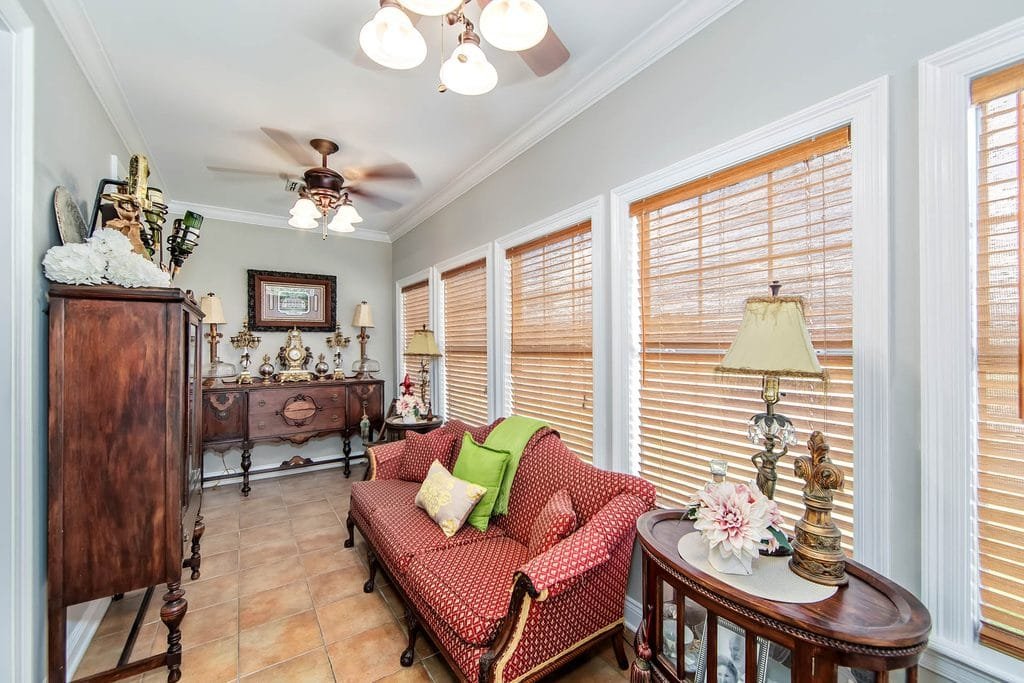 A sunroom with large windows, blinds, a red patterned loveseat with throw pillows, antique furniture, decorative lamps, and tile flooring under ceiling fans with lights—perfectly showcasing classic design touches often highlighted in realtor photography examples.