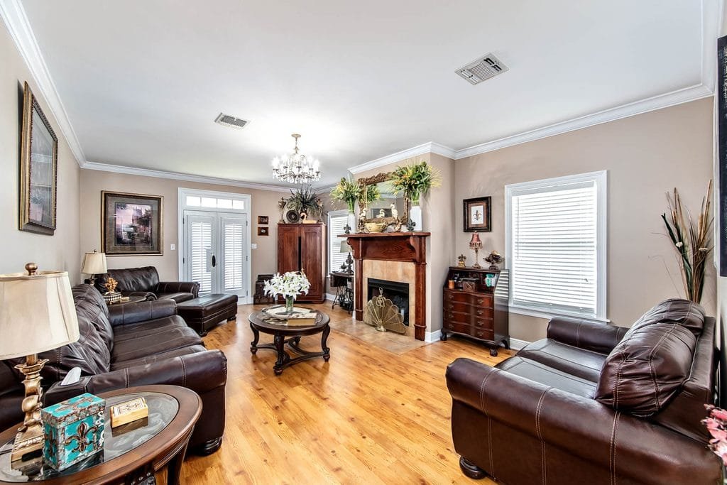 Bright living room with brown leather sofas, wooden floor, coffee table, chandelier, fireplace with decor, framed art, large windows, and light beige walls. Green plants and a ceiling vent add detail—an inviting space that stands out in realtor photography examples.