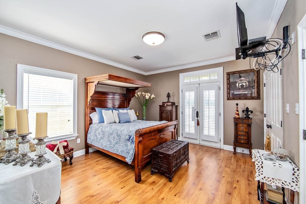 Elegant bedroom with a wooden canopy bed, blue and white bedding, hardwood floors, a trunk at the bed’s foot, two windows, French doors, and various decorative furniture pieces and accessories—an inviting space that perfectly showcases realtor photography examples.