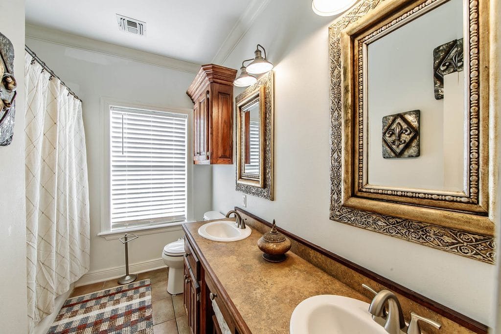 A bathroom with two sinks, large framed mirrors, wooden cabinets, a toilet, a shower with a white curtain, a window with blinds, and wall-mounted lights above the mirrors—an inviting space that highlights classic features often showcased in realtor photography examples.