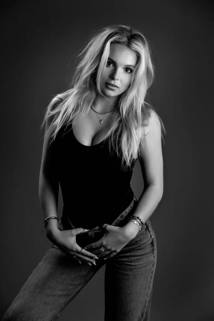 Black and white photo of a woman with long hair, wearing a sleeveless top and jeans, posing with her hands resting on her waist, looking directly at the camera against a dark background—an ideal shot for model portfolio photos for agencies.