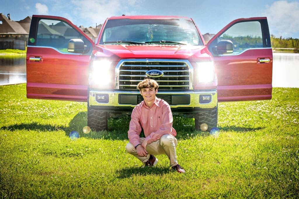 CooperC_07_web A young man in a pink shirt and khakis kneels on grass in front of a red Ford truck with open doors and headlights on, capturing the perfect senior pictures with cars. Houses and a lake provide a scenic backdrop for this memorable photo.