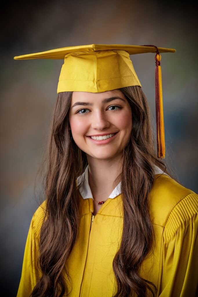 Young woman wearing a yellow graduation cap and gown smiles at the camera against a blurred, neutral background—a perfect example of the joyful moments you’ll want to capture when deciding when to book senior pictures in South Louisiana.