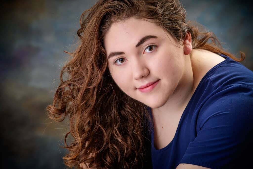 A person with long, curly brown hair and fair skin smiles slightly, wearing a blue shirt in this senior portrait. The background is softly blurred with blue and brown tones. Houma Photographer Kirk Voclain Photography