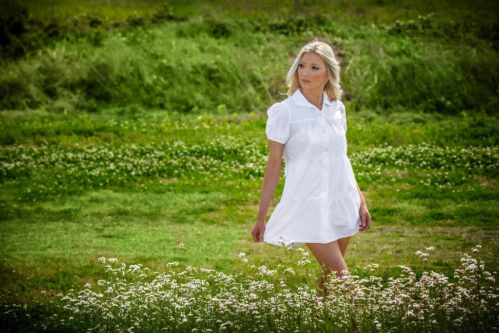A woman in a short white dress stands in a field of green grass and small white flowers, looking to the side on a sunny day—one of the best senior picture locations in Thibodaux, LA.