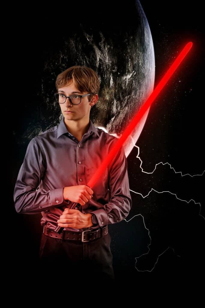A camera-shy high school senior poses confidently in glasses, holding a glowing red lightsaber against a dark space background with a planet and outlines of continents behind them—a unique twist on traditional senior portraits in Houma.