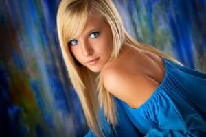 A young woman with long blonde hair and bright blue eyes poses in a blue off-the-shoulder top against a colorful, blurred background with blue and green tones—an inspiring example for those looking to choose senior portrait colors that beautifully highlight natural features. Kirk Voclain Photography Houma Photographer