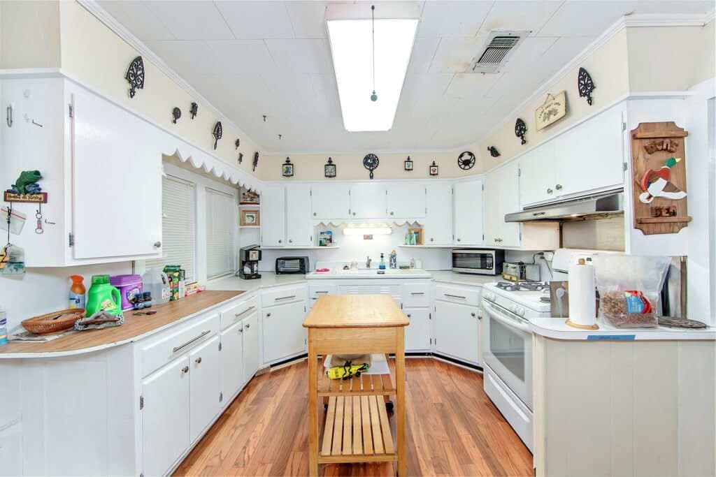 Bright kitchen with white cabinets, wooden island, and various appliances. Decorative wall clocks and knick-knacks adorn the space. Wooden floors and a fluorescent ceiling light complete the room—showcasing why hire property photographer professionals to truly capture this inviting atmosphere.