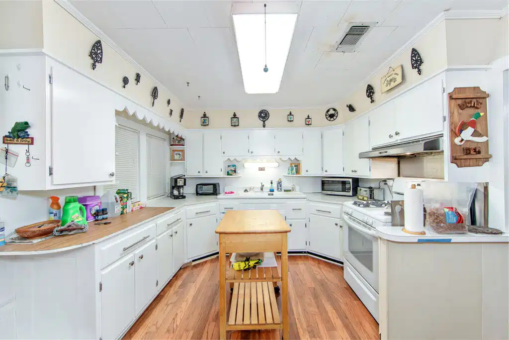 Bright kitchen with white cabinets, wooden island, and various appliances. Decorative wall clocks and knick-knacks adorn the space. Wooden floors and a fluorescent ceiling light complete the room—showcasing why hire property photographer professionals to truly capture this inviting atmosphere.