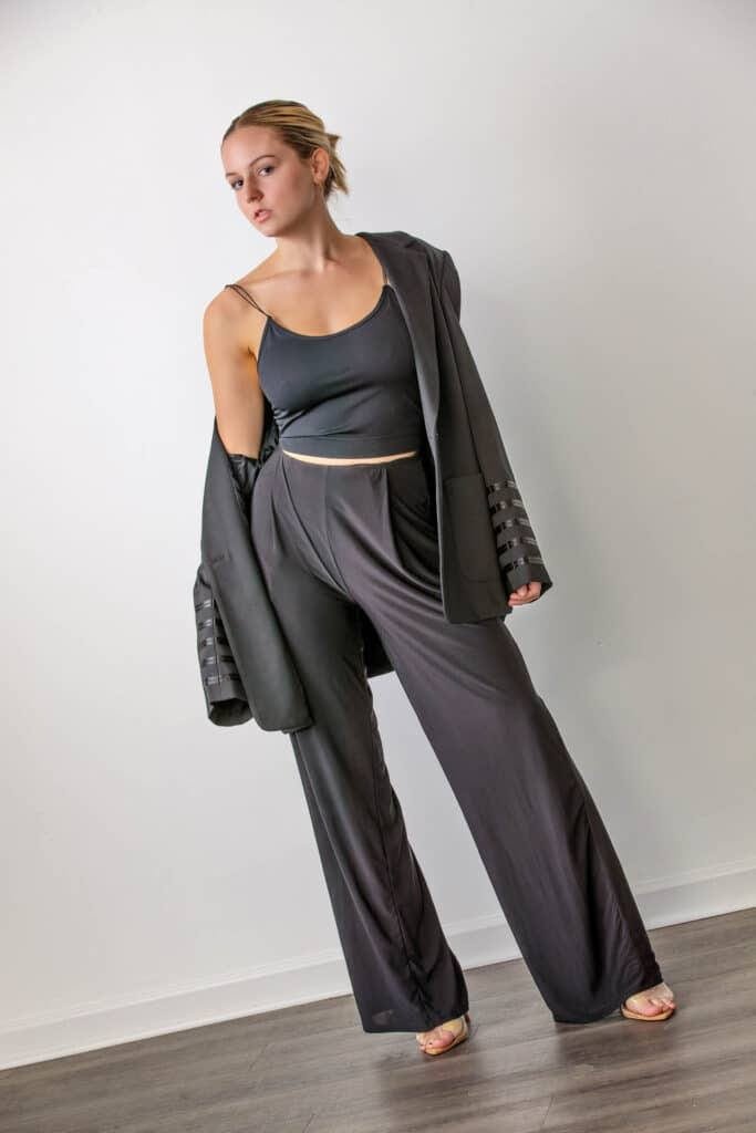 Young woman poses in a black crop top, high-waisted wide-leg pants, and an oversized blazer draped over her shoulders, standing against a plain white wall on a wooden floor—perfect inspiration if you want to choose senior portrait accessories that add modern elegance to your look.