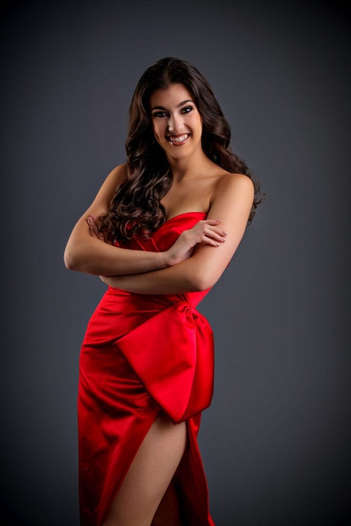 Woman with long dark hair wearing a strapless red dress stands against a dark gray background, smiling with arms crossed and one leg slightly showing through a slit in the dress, effortlessly demonstrating natural posing versus stiff poses.
