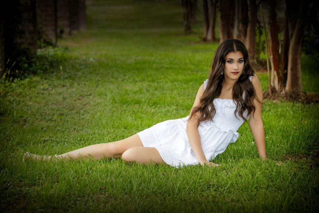 A woman with long brown hair wearing a white dress sits on green grass in an outdoor setting, looking at the camera with a relaxed expression—perfect inspiration if you want to plan a relaxed portrait session. Trees and greenery are visible in the background.