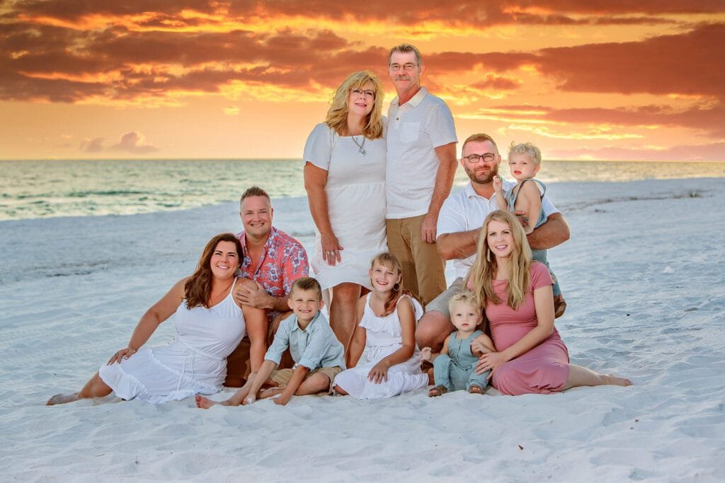 Three adults stand behind six adults and children sitting and kneeling on white sand at the beach, with a vibrant orange sunset and ocean in the background. Everyone is dressed in light, summery clothing and smiling at the camera, perfectly capturing a moment that answers all your family portrait questions about joy, togetherness, and natural beauty. Kirk Voclain Houma Photographer.