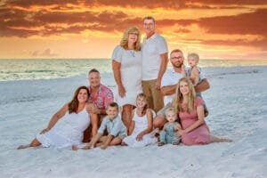 Three adults stand behind six adults and children sitting and kneeling on white sand at the beach, with a vibrant orange sunset and ocean in the background. Everyone is dressed in light, summery clothing and smiling at the camera, perfectly capturing a moment that answers all your family portrait questions about joy, togetherness, and natural beauty. Kirk Voclain Houma Photographer.