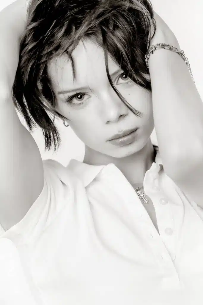 A close-up black and white natural portrait of a person with short hair pulling their hands up to their head, wearing a collared polo shirt and a cross necklace.